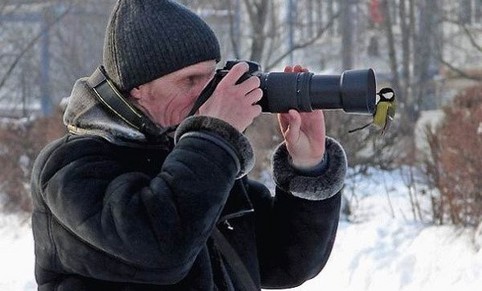 Ein Mann schaut durch einen Fotoapparat, auf das Objektiv hat sich ein Vogel gesetzt.